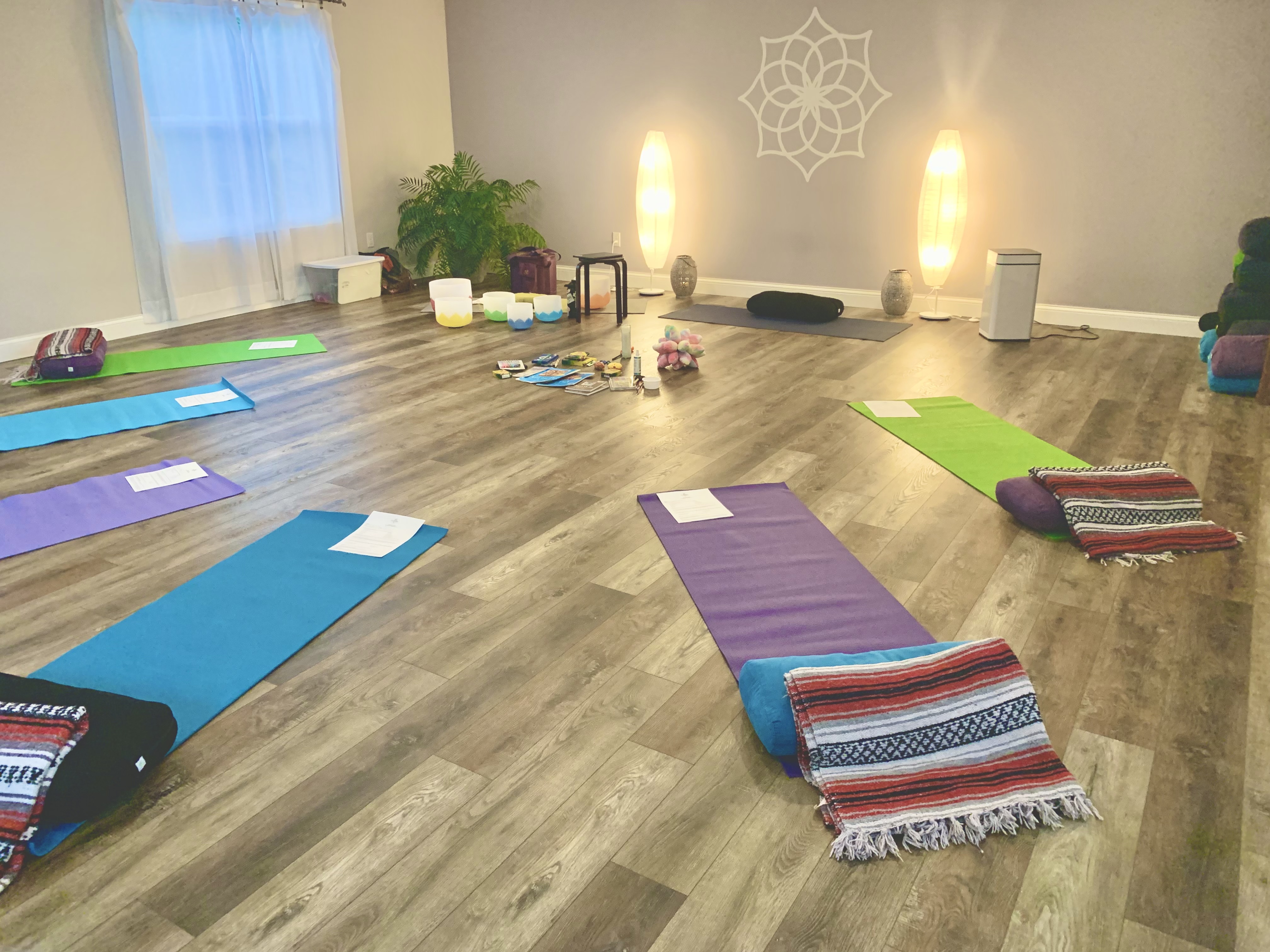 Image of a somatics studio with 6 mats set out in colors of blue, green and purple, blankets near each mat, sound bowls in the corner, a palm plant and two tall lanterns, all atop a grey wood floor with grey walls