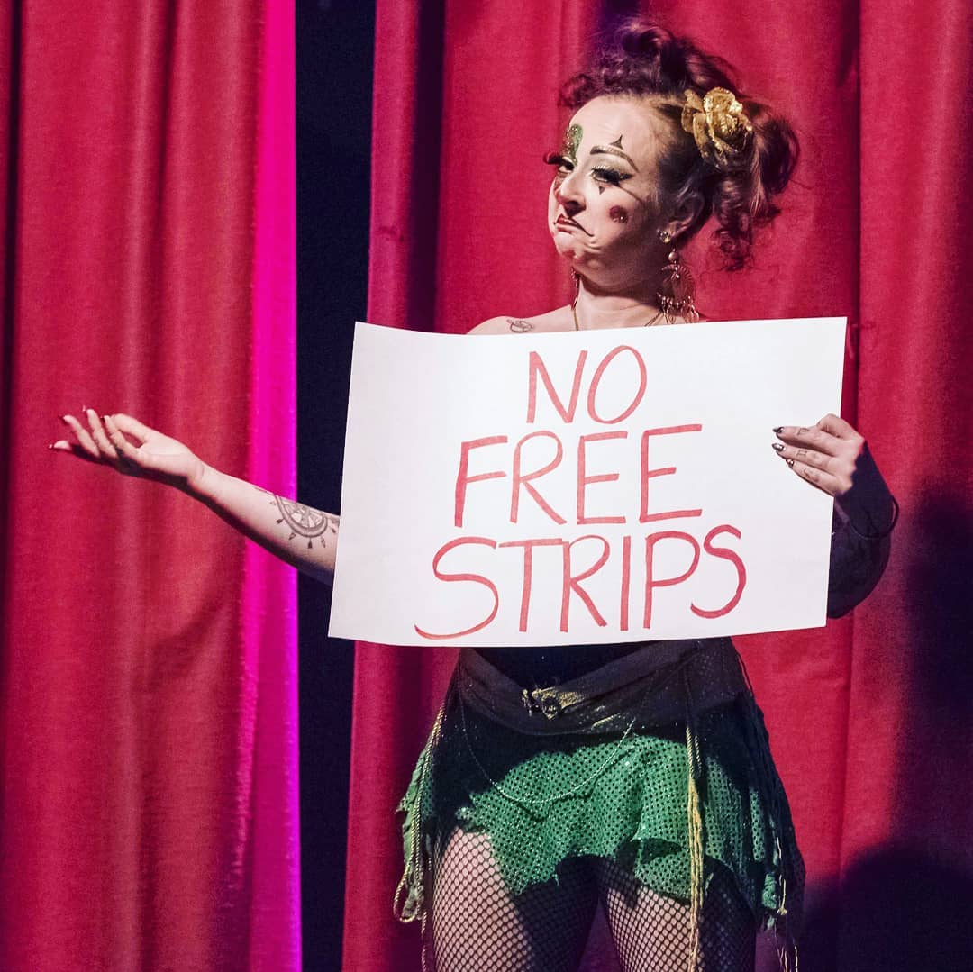 Photo of Deanna dressed as a clown with a green sequin skirt, fishnets, red hair in an updo and are holding a white sign with red lettering that says “no free strips” in front of a red theatre curtain.