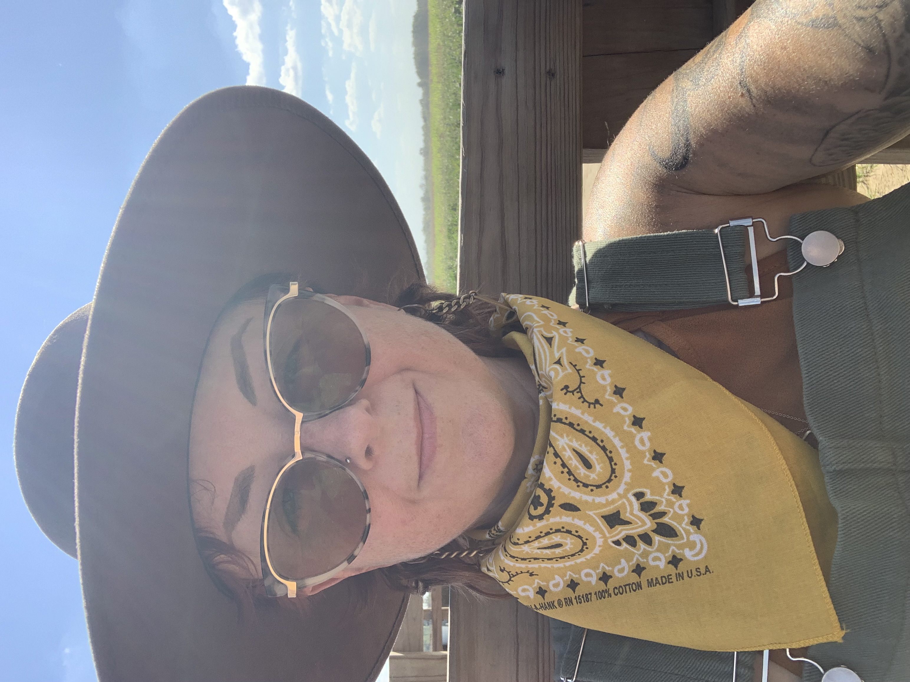 Photo of Deanna Sophia Danger, they are wearing a wide brim brown hat, overalls, yellow bandana around their neck and tinted glasses. They are sitting in front of a wooden railing with grass & sky in the background