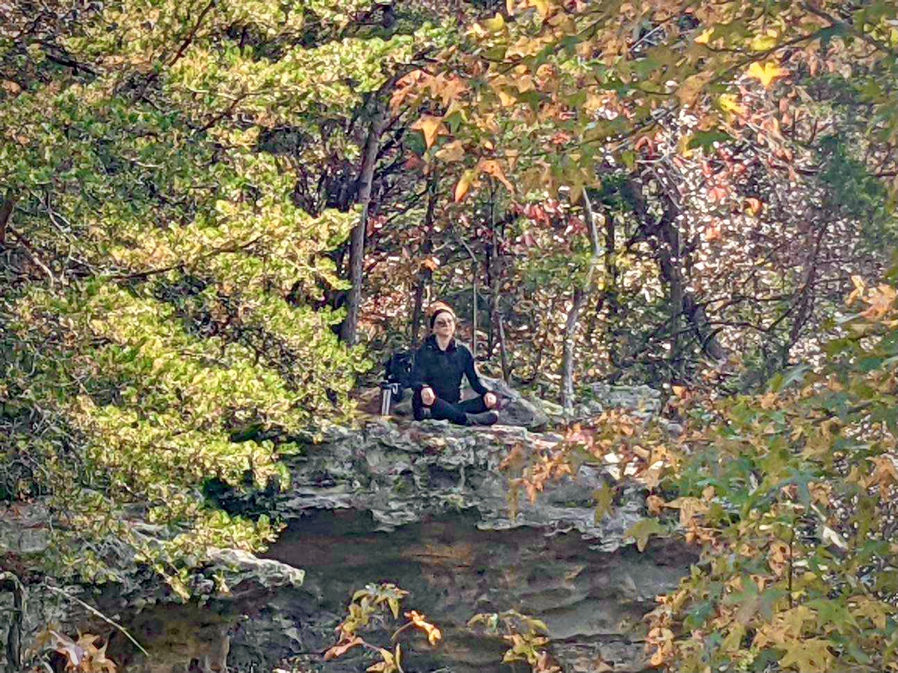 Deanna wears all black and sits meditating atop a rocky outcropping amidst green and fall leaves