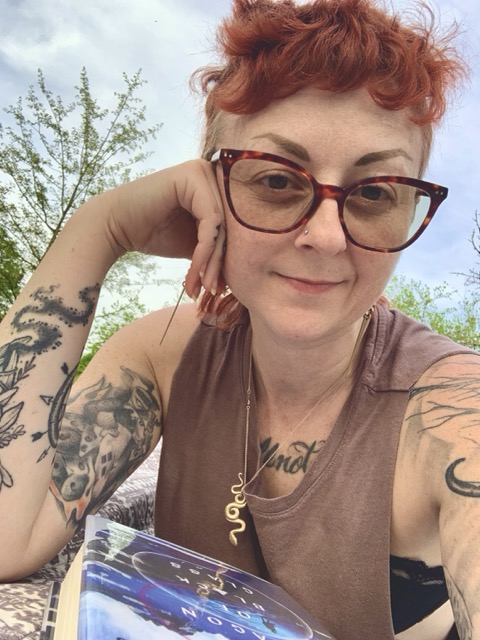 Photo of Deanna with short red hair, red frame glasses, a brown shirt, tattoos on their arms and chest, a slight smile on their face. They are laying in a park with a book in front of them and green trees in the background.